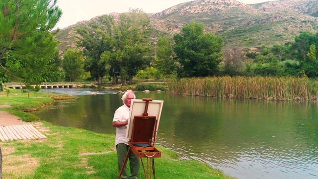 一位上了年纪的艺术家在秋天的河边的画架上画一幅画视频下载