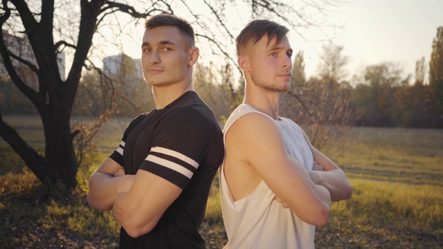 两个年轻的白人健美运动员站在阳光下看着镜头。英俊强壮的运动员花时间在秋天公园。健康的生活方式，运动的理念。视频素材
