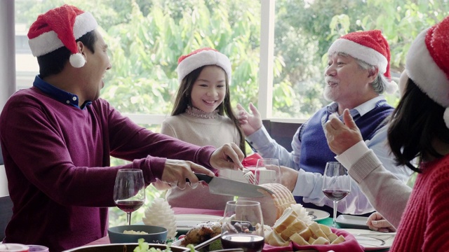 亚洲家庭正在吃圣诞晚餐，爷爷、奶奶、父母和女儿们愉快地坐在一起聊天。爷爷收到了一份礼物。人们戴着红色的圣诞老人帽。新年的庆祝活动视频素材