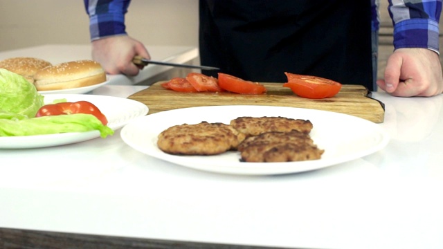 一个穿着围裙的男人在厨房里准备一个汉堡，桌子上是奶酪汉堡的原料和切片西红柿，慢动作视频素材