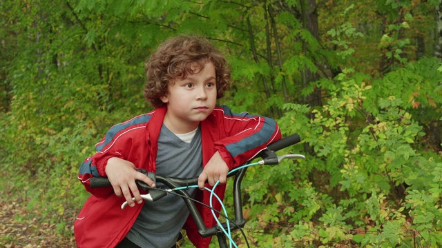 在夏日公园里的卷发男孩。近距离面对十几岁的男孩在绿色公园骑自行车。在绿色公园英俊的男孩看着镜头。视频下载