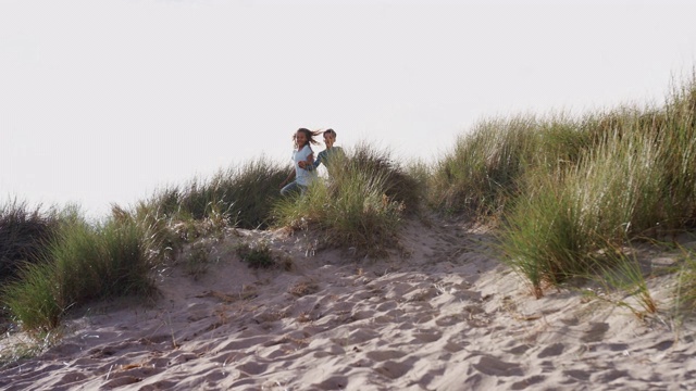 两个孩子在冬季海滩度假的沙丘上奔跑跳跃-用慢镜头拍摄视频素材