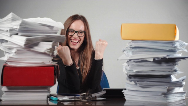 微笑的女办公室经理显示幸福的情绪坐在两堆完成的文件。成功的总经理在镜头前举手示意，心情愉悦。过度工作的上班族概念视频素材