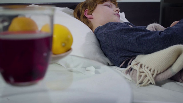 一个白人小男孩躺在床上，背对着镜头。前台的桌子上放着热饮和橘子。医疗、疾病、疾病。视频素材