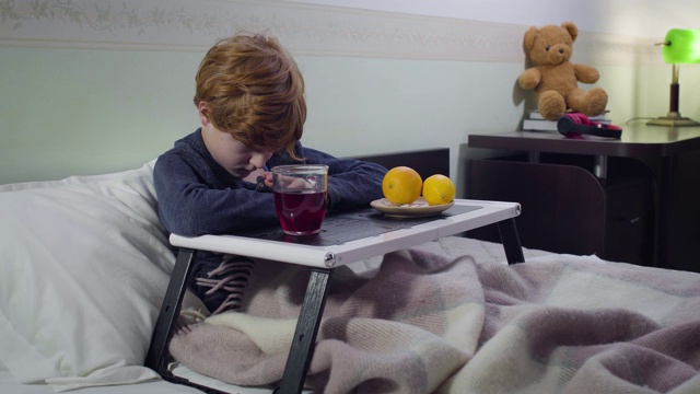 心烦意乱的白人男孩坐在床上，托盘上放着热饮和橘子。一个伤心可爱的红发小孩呆在家里。医疗、医药、疾病。视频素材