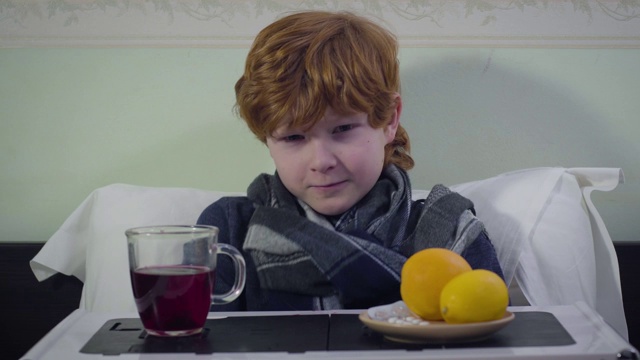红发白人男孩打喷嚏，看着镜头。生病的红发小孩坐在家里的床上。医疗、疾病、药物。视频素材