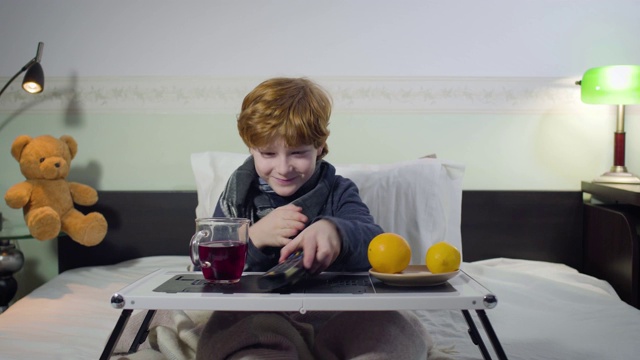 病态白人男孩用遥控器切换频道看电视的肖像。可爱的红发小孩坐在床上，面前放着茶和橘子。疾病、医疗保健、生活方式。视频下载