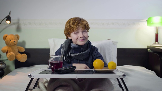 开朗的红发白人男孩一边看电视一边笑。可爱的生病的孩子坐在床上，托盘上有遥控器，茶和橘子。生活方式、休闲、医疗保健。视频素材