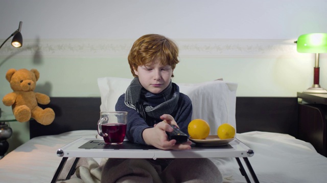咳嗽的白人小男孩拿着遥控器切换电视频道。红发可爱的孩子坐在床前，托盘上放着茶和橘子。疾病、健康的生活方式。视频素材