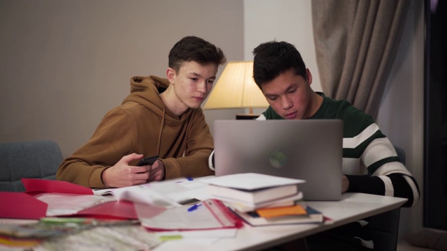 注意力集中的亚洲少年向正在使用智能手机的白人同伴展示笔记本电脑屏幕。两个英俊的男大学生在网上学习。远程教育，生活方式，现代科技。视频下载