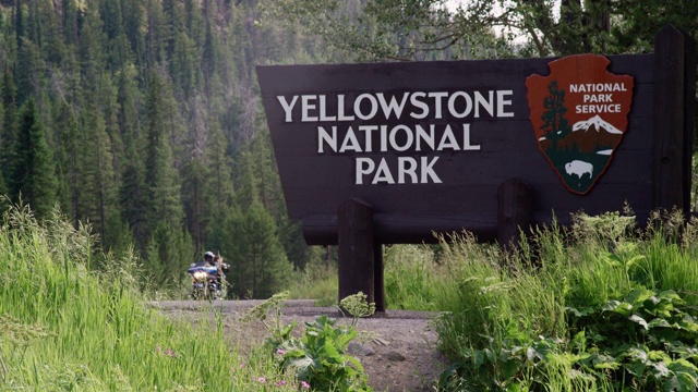 一辆吉普车和一辆摩托车行驶在黄石国家公园的欢迎标志旁，周围是森林和落基山脉视频下载