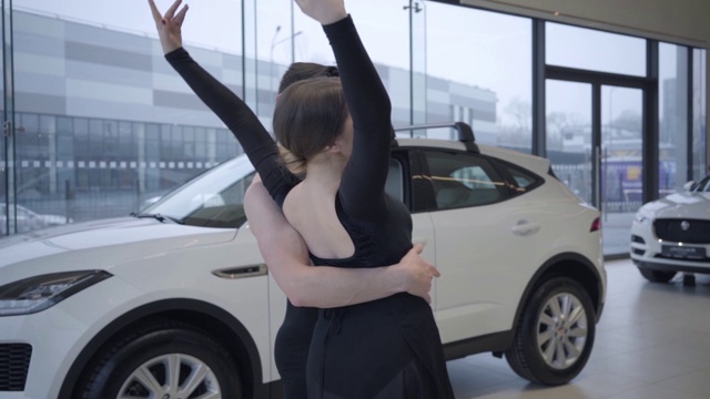 慢动作的微笑白人妇女弯背支持的男人。快乐的芭蕾舞演员在汽车经销店跳舞。汽车工业，高雅，艺术，美丽。缓慢的运动。视频下载