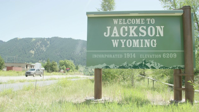 在一个阳光明媚的日子里，汽车和车辆驶过“欢迎来到怀俄明州杰克逊”的标牌，标牌的背景是群山视频素材