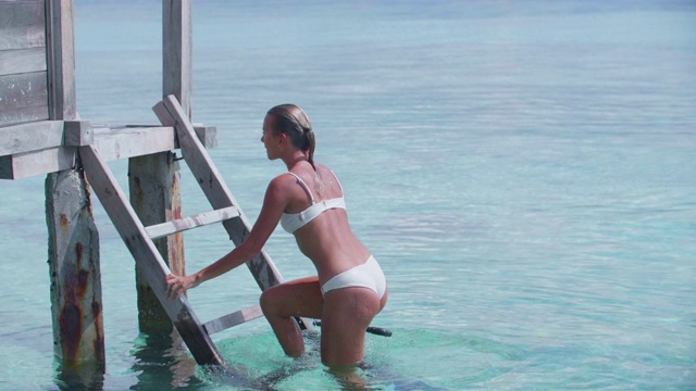 一个穿着白色比基尼的女人在一个热带岛屿酒店度假时，穿着潜水面罩的泳衣爬梯子。视频素材
