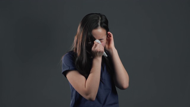 一个年轻的亚洲女孩用纸巾打喷嚏。孤立的女人在灰色工作室的背景是生病，有感冒或过敏反应。冠状病毒，2020年流行，疾病概念。视频下载