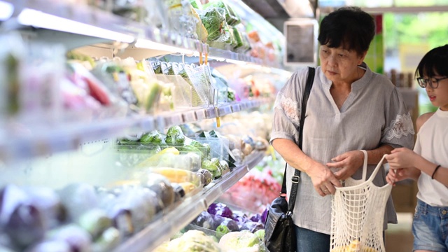 一位亚洲华人的祖母和孙女在杂货店的冰箱蔬菜区挑选和购买蔬菜视频下载