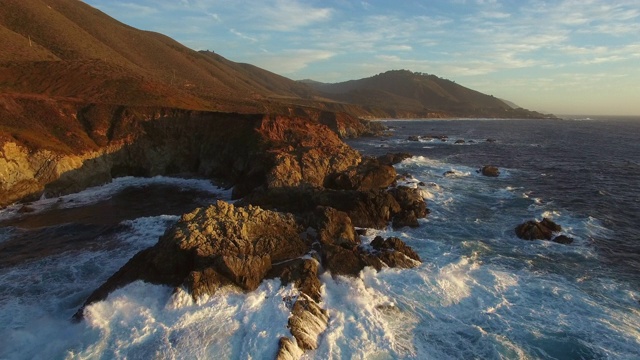 航拍:无人机接近岩石海岸，日落时海浪在海滩上溅起浪花，天空映衬下的山景——大苏尔，加利福尼亚视频下载