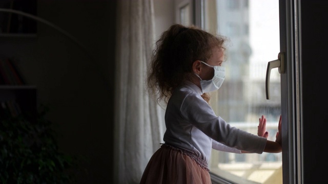 2019冠状病毒病概念，小女孩戴着口罩透过窗户往外看，带着纸条隔离在家。可怜的孩子因为冠状病毒被隔离。视频下载