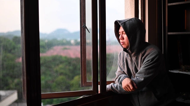 日落时分，一名身穿连帽衫的亚洲华人中年男子从家中望向窗外，表情严肃视频素材