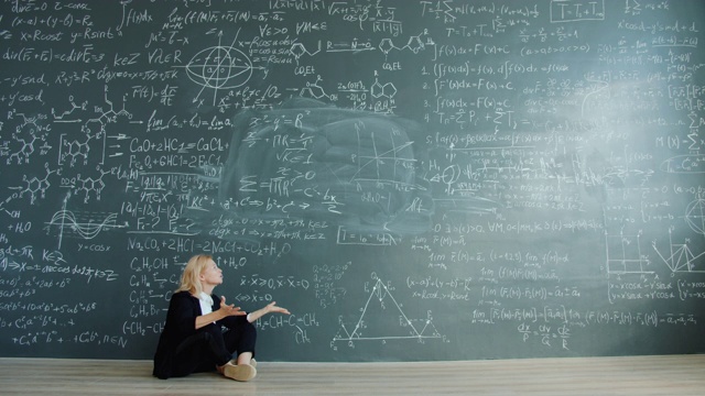 一位困惑的女科学家坐在黑板旁的地板上，用手势说话视频素材