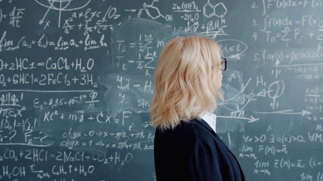 在大学教室里，一个金发成人走到黑板前，上面写着科学公式视频素材