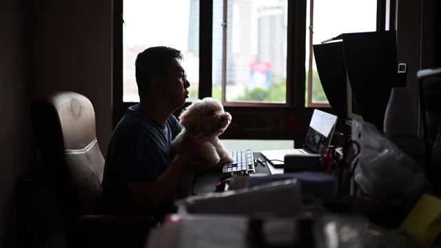 一位亚洲华人中年男子在网上与朋友、家人和商业伙伴聊天、聊天，一边在家里办公室的电脑屏幕上看着摄像头聊天，一边带着他的玩具狮子狗视频下载
