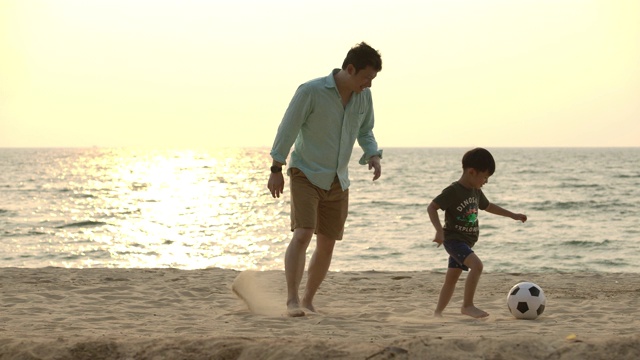 亚洲父亲和儿子一起在沙滩上玩足球在夕阳的背景下。缓慢的运动。幸福的家庭的概念。视频下载