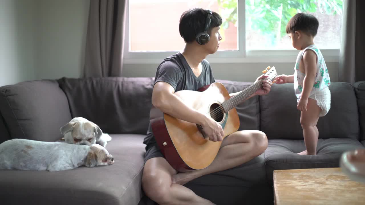 父亲和蹒跚学步的男孩在家里的客厅里弹吉他。视频下载