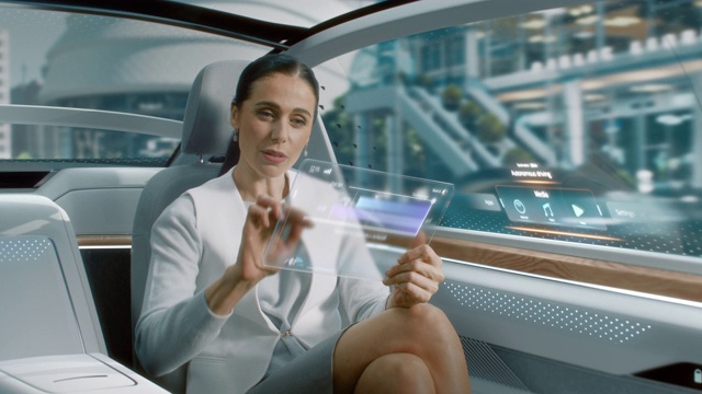 迷人的资深女性坐在自动驾驶汽车的后座上，在未来透明平板电脑上阅读新闻与增强现实界面。自动驾驶货车在公共道路上行驶。视频素材