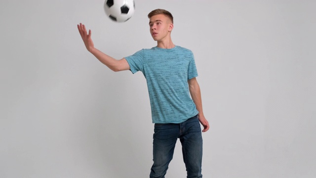一个十几岁的男孩站在足球的肖像，用他的脚扔它，他微笑的脸在镜头前的白色背景慢镜头。足球球迷,球员。青少年和体育视频下载
