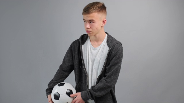肖像少年严肃地旋转着不耐烦地把足球从一只手扔到另一只手，看着灰色背景的摄像机在电视上观看比赛。青少年和运动。生活方式视频素材