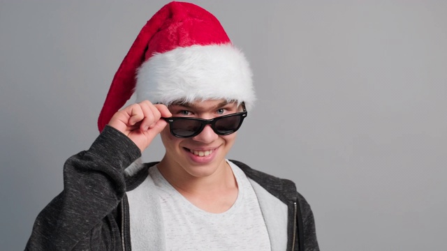 快乐的少年男孩在圣诞帽进入框，举起太阳镜微笑指着他的手指在灰色的背景相机。诺埃尔。副本的空间。圣诞情绪，传统情感。冬天。诺埃尔视频素材