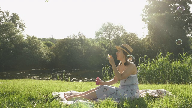 戴着帽子、穿着衣服的快乐微笑的女人在公园里吹泡泡，背景是河流和树木。可爱的年轻女士坐在公园草地上吹泡泡，高兴和兴奋视频素材