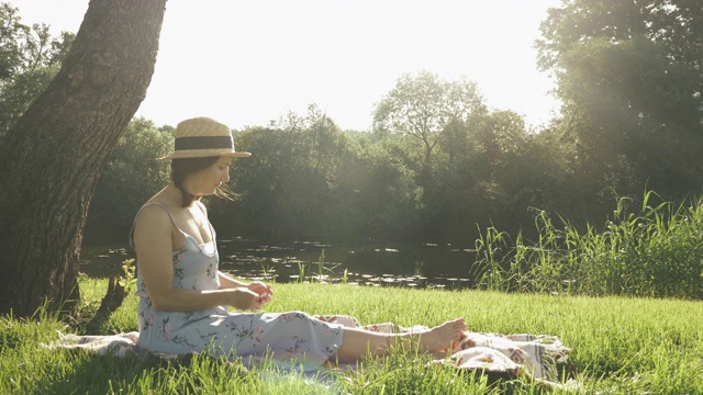 女孩戴着草帽，穿着衣服，坐在毯子上，摘下耳机，在公园里放松，享受阳光明媚的夏日。可爱迷人的快乐女士周末在公园的河边。女性在公园休息视频素材
