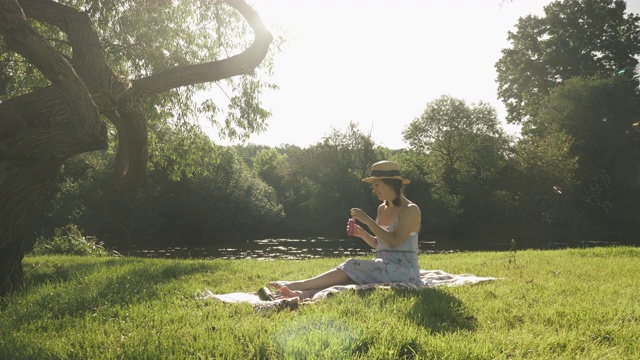 时髦快乐的女人在河边的公园吹泡泡。年轻的黑发女孩坐在河边的草地上，在阳光明媚的日子里放松和吹泡泡。在公园里享受暑假的女士视频素材