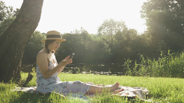 一名女子戴上耳机，坐在公园大树旁的格子布上，以河流为背景听音乐。年轻快乐的女性听着音乐，享受着温暖阳光的周末户外视频素材