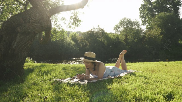 女人在公园里放松，戴着耳机听着音乐，享受着暑假。可爱迷人的女孩戴着帽子和衣服躺在毯子上在河边的草地上放松视频素材