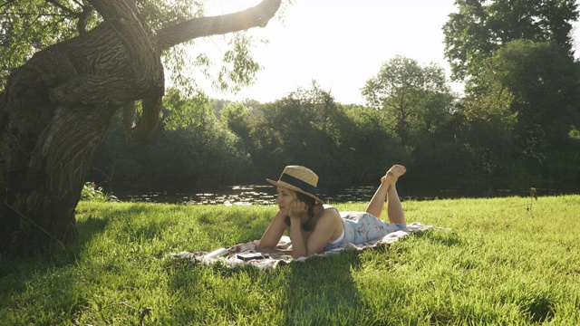 戴着帽子，盘腿躺在河边大树下的公园草地上，悠闲地享受着温暖的阳光。年轻女子花时间在公园附近的河在暑假视频素材
