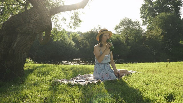 穿着草帽的妇女坐在河边公园草地上的毯子上，喝着瓶子里的水。女士在阳光明媚的夏日户外放松。女性坐在大自然的草地上视频素材