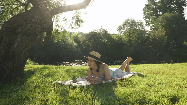 女孩在河边的草地上休息，享受着温暖的阳光。漂亮的黑发女子戴着草帽躺在河边的大树下的草地上，阅读日记和放松的户外视频素材