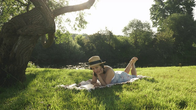 戴帽子的女人躺在草地上，以河流为背景，在大自然中读书。年轻迷人的女士穿着小蓝衣服在公园的草地上放松，靠近大树，享受暑假视频素材