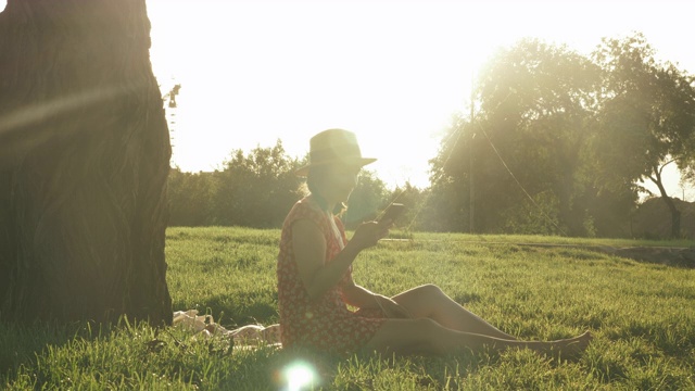 微笑着快乐的女士听着耳机里的音乐，看着夕阳，在公园里享受着温暖的夏夜。在公园里享受周末的女人。一个女人坐在一棵大树旁的草地上视频素材