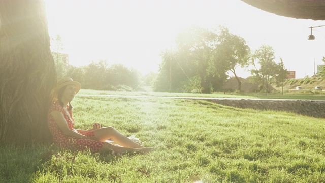 身着红色礼服，头戴草帽，坐在草地上，倚着公园里的一棵大树，休息着，吃着苹果。戴着帽子的女人享受周末假期。穿着休闲的女性视频素材