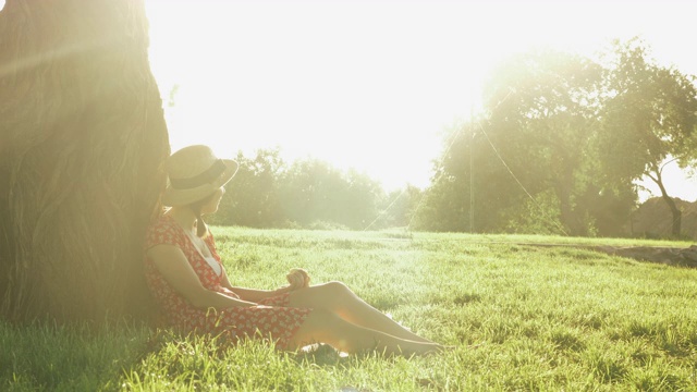 一个女人坐在公园的草地上，在一个阳光明媚的夏天的晚上。年轻迷人的女士，戴着草帽，穿着衣服，倚在一棵大树上，坐在公园的草地上，放松和欣赏美丽的日落视频素材