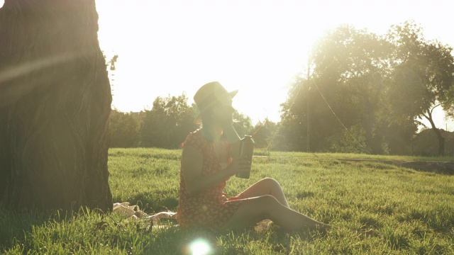 戴着帽子的迷人女士坐在公园的格子上，在温暖的夏夜喝着瓶子里的水，放松和欣赏美丽的日落。穿着衣服的女人在公园里放松。女孩周末在公园视频素材