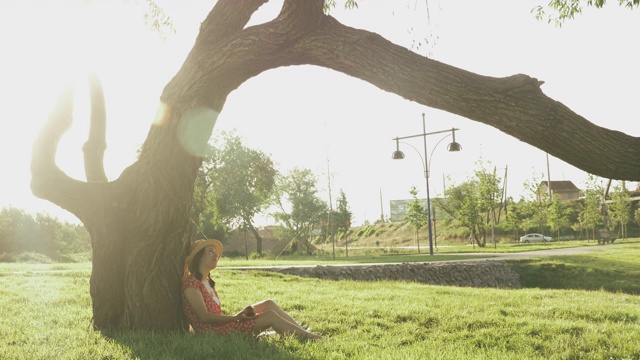快乐的年轻女子休息在公园附近的大树和吃苹果。可爱的女性放松在公园日落。戴草帽的迷人女孩坐在公园的草地上，享受夏日的夜晚视频素材