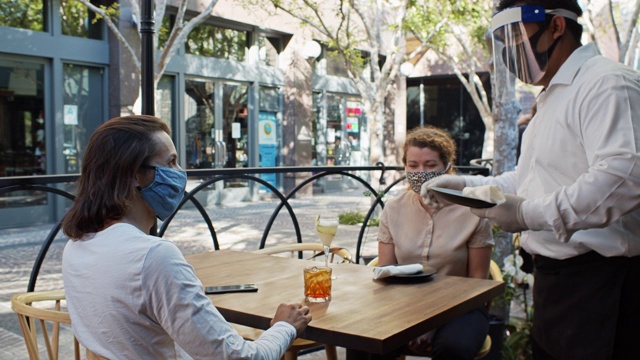 2019冠状病毒病大流行期间，穿着个人防护用品的服务员为户外餐桌上的顾客提供餐具视频下载