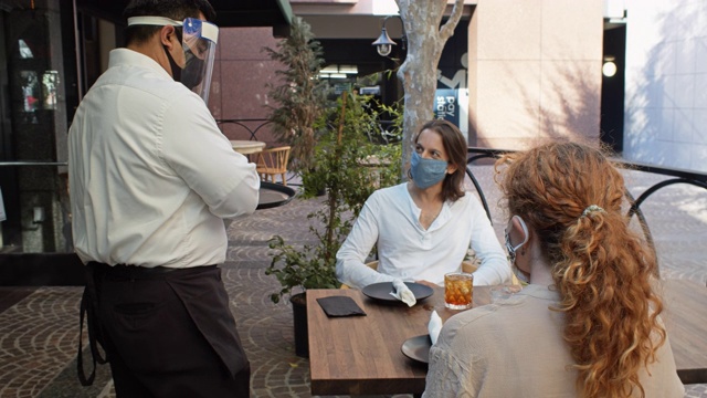 在新冠肺炎大流行期间，穿着个人防护用品的服务员在户外餐桌为蒙面顾客服务视频下载