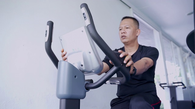 一名40-50岁的亚洲男子正在健身房里骑电动自行车减肥。视频下载
