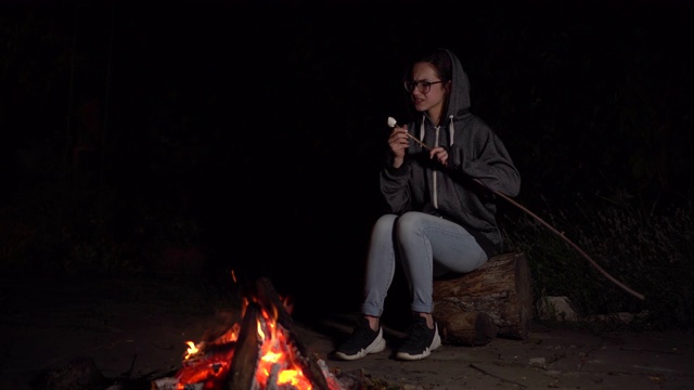 一个戴兜帽的年轻女子在火上烤棉花糖，棉花糖就着火了。晚上篝火。视频素材